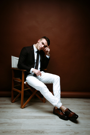 Portrait of handsome stylish man in elegant black suit and white pants sitting on chairの写真素材
