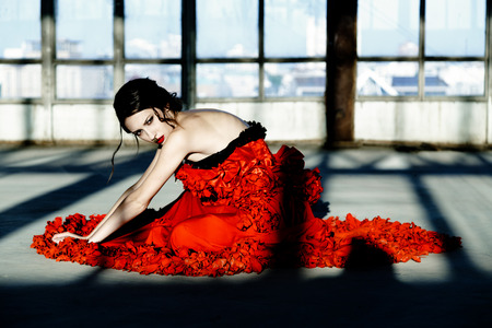 Beautiful, brunette woman in an old, abandoned house and sitting on floor, wearing a red dress,の写真素材
