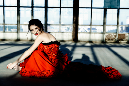 Beautiful, brunette woman in an old, abandoned house and sitting on floor, wearing a red dress,の写真素材
