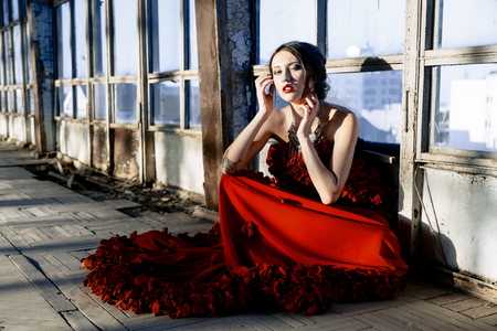 Beautiful, brunette woman in an old, abandoned house and sitting on floor, wearing a red dress,の写真素材