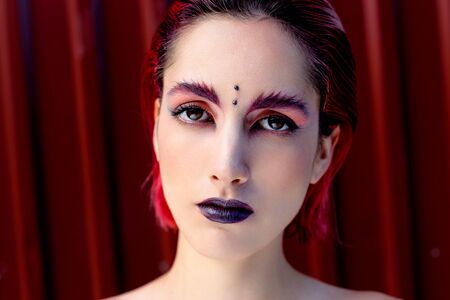 Attractive young woman with colorful makeup, red hairstyle, expressive eyes and piercing on forehead, red metal wall on backgroundの写真素材