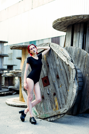 Attractive young woman with colorful makeup and red hairstyle in simple black bodywear sitting on big wooden reelsの写真素材