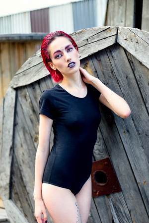Attractive young woman with colorful makeup and red hairstyle in simple black bodywear sitting on big wooden reelsの写真素材