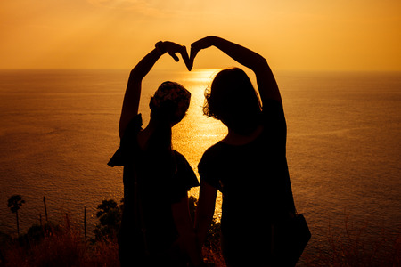Silhouette photo of two girls arranged their arms in heart shape with sunset backgroundの写真素材