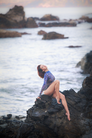 gymnast girl sitting on a rock with bare legs and back in an acrobatic tights against the seaの写真素材