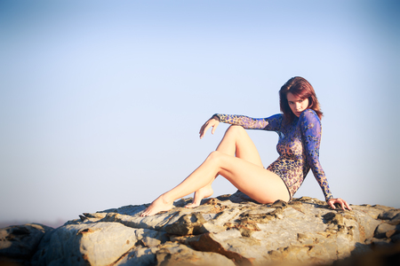 brunet gymnast girl sitting at a rock with bare legs and back in an acrobatic tights against seaの写真素材