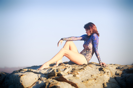 brunet gymnast girl sitting at a rock with bare legs and back in an acrobatic tights against seaの写真素材