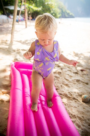 small blonde baby girl in swim suit walk on pink mattress on the beachの写真素材