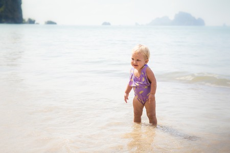small blonde baby girl in purple swim suit play with waves on seashoreの写真素材