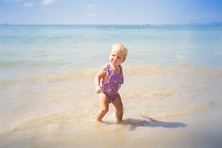 small blonde baby girl in purple swim suit play with waves on seashoreの写真素材