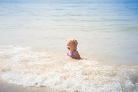 small blonde baby girl in purple swim suit play with waves on seashoreの写真素材