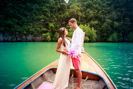 tall handsome groom hold his young brunette bride in long wedding dress stand on longtail boat in green lagoon among cliff islandの写真素材