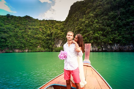 brunette bride in long white wedding dress hugging her young handsome groom standing on longtail boat in thai island lagoonの写真素材