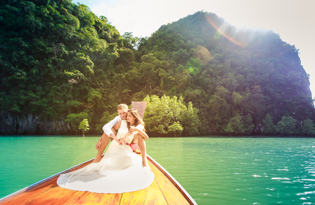 handsome groom hugging his brunette bride in white wedding dress sitting on longtail boat in thai island lagoonの写真素材