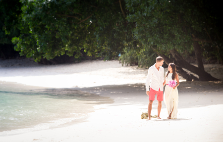 handsome groom walk with pretty brunette bride in long white wedding dress on island beach white sand in Thailandの写真素材