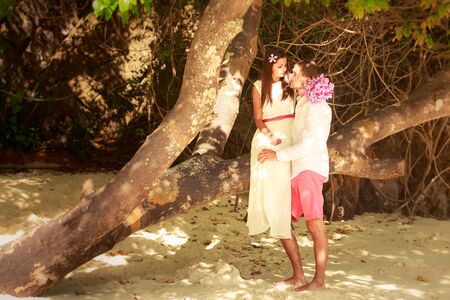 brunette bride in long white wedding dress sitting on the big tree and hold her handsome groom on island beach in Thailandの写真素材