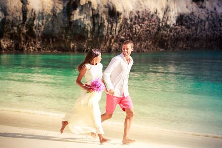 handsome groom run with his beautiful brunette bride in long white wedding dress on island bay with green mountain background in Thailandの写真素材