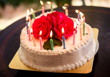 candle burning on a cream white creamy delicious cake decorated with three red roses on the topの写真素材