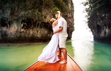 blonde bride kissing and embracing strong handsome groom standing on longtail boat against cliff islandの写真素材