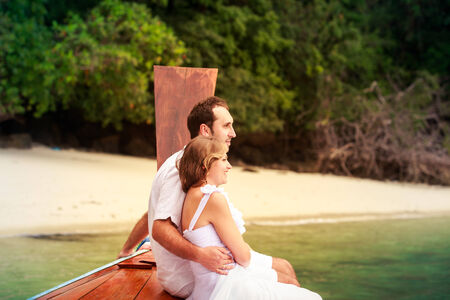 handsome groom embrace blonde bride in wedding dress sitting on longtail boat near the rocky islandの写真素材