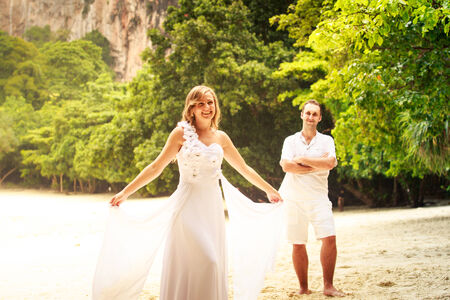 blonde bride and handsome groom stand on the sand beach barefoot near green jungle treesの写真素材