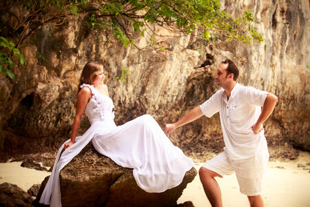 handsome groom looks at beautiful bride sitting on the rock at the sand beachの写真素材