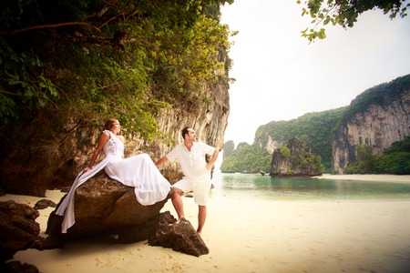 handsome groom looks at beautiful bride sitting on the rock at the sand beachの写真素材