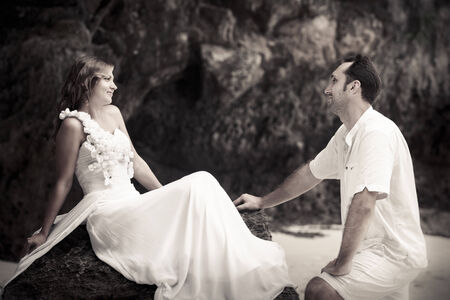 handsome groom looks at beautiful bride sitting on the rock at the sand beachの写真素材