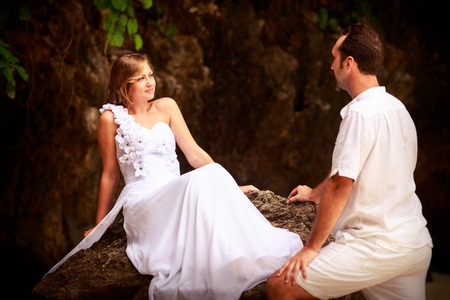 handsome groom looks at beautiful bride sitting on the rock at the sand beachの写真素材