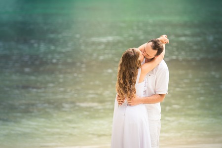 pretty blonde bride and handsome groom kiss standing at sand beach against azure sea backgroundの写真素材