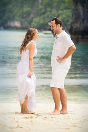 pretty blonde bride and handsome groom talk standing at sand beach against azure sea backgroundの写真素材