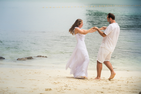 young blonde bride and handsome groom dance at sandy beach against azure sea backgroundの写真素材