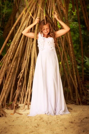 young blonde bride in white wedding smiling against strange roots of tropical plantの写真素材