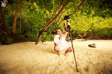 young handsome groom in white holds camera on sand beach against tropical plantsの写真素材