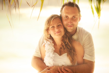handsome groom smile and embraces blonde bride under the palm with azure sea on backgroundの写真素材