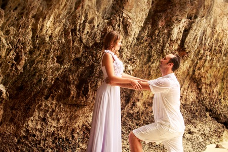handsome groom stands on his knees and looks at blonde bride against rock at sand beachの写真素材