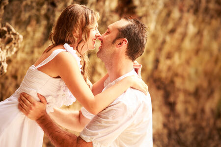 handsome groom stands on his knees and kiss blonde bride against rock at sand beachの写真素材