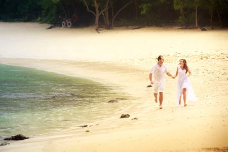 blonde bride and handsome groom run barefoot on sandy island beach against rocks and water backgroundの写真素材