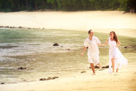 blonde bride and handsome groom run barefoot on sandy island beach against rocks and water backgroundの写真素材