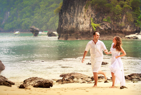blonde bride and handsome groom run barefoot on sandy island beach against rocks and water backgroundの写真素材