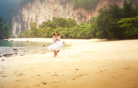 blonde bride and handsome groom whirl and kiss on sandy beach against green cliff and azure sea backgroundの写真素材