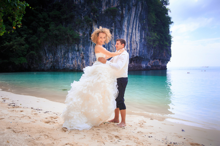 handsome groom raise up his blonde bride in elegant wedding dress near cliff on island beach Thailandの写真素材