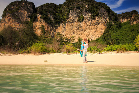 handsome man hold his beautiful blonde wife in elegant dress on island beach with green mountain on backgroundの写真素材