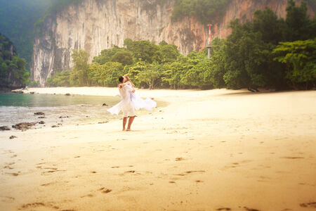 blonde bride and young groom whirling on sandy beach against jungle cliff rocks and water backgroundの写真素材