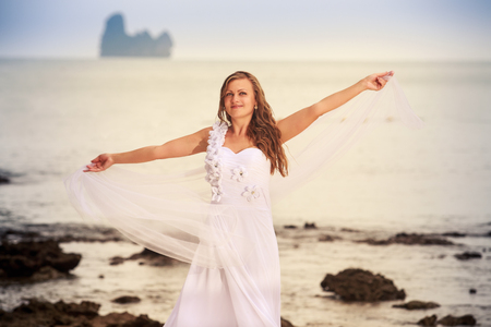 blonde bride in wedding dress whirls with white veil stand barefoot on a sand beach against cliff and island sea water backgroundの写真素材