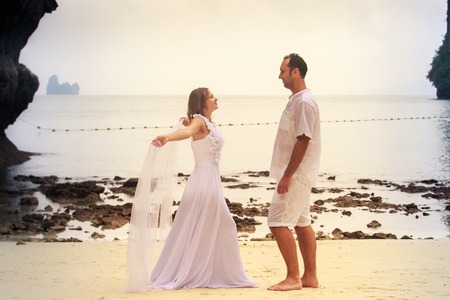 blonde bride and young groom dance barefoot with white veil standing on sandy beach against cliff and island sea water backgroundの写真素材