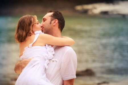 young groom kiss his blonde bride standing on sandy beach against cliff and island sea water backgroundの写真素材