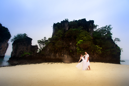 young groom hold his blonde bride standing on sandy beach against cliff and island sea water backgroundの写真素材