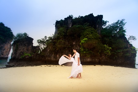 young groom hold his blonde bride standing on sandy beach against cliff and island sea water backgroundの写真素材