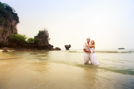 young groom hold his blonde bride standing at sea waves against cliff islandの写真素材